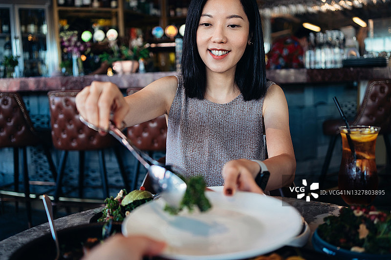 年轻的亚洲夫妇在餐厅享受晚餐约会。微笑的年轻女子提供新鲜的绿色沙拉在盘子里，并传递给对面桌子上的男友。外出就餐的生活方式图片素材