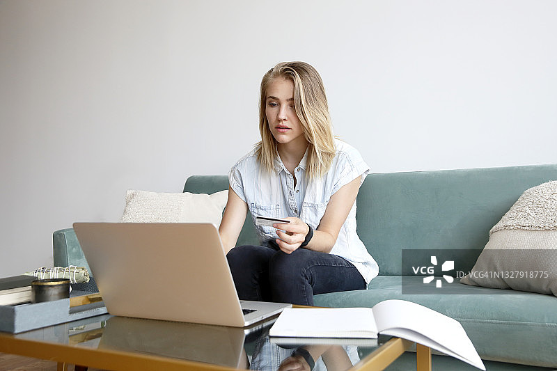 一名金发的白人千禧女性穿着休闲装，在家里用笔记本电脑支付信用卡。图片素材