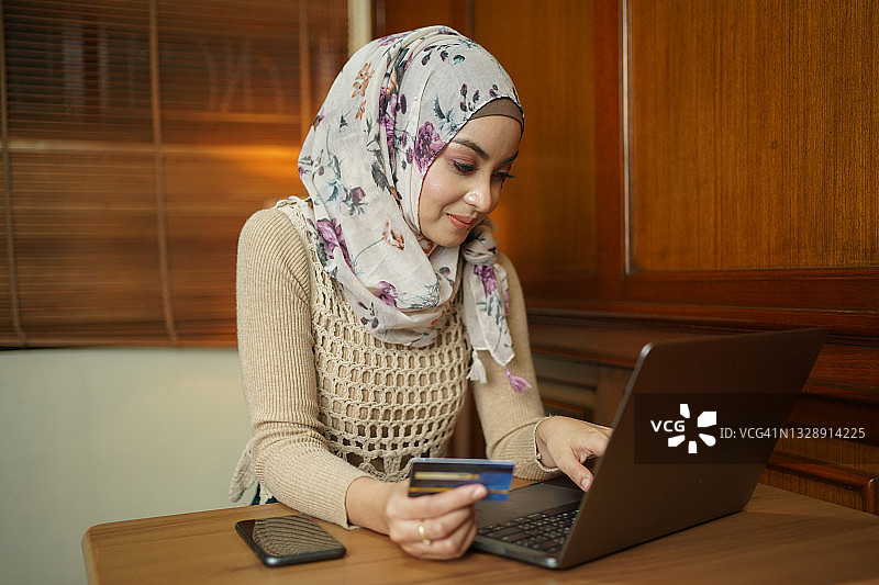亚洲穆斯林妇女持有信用卡，使用笔记本电脑进行网上购物和网上支付通过互联网。技术的概念。图片素材