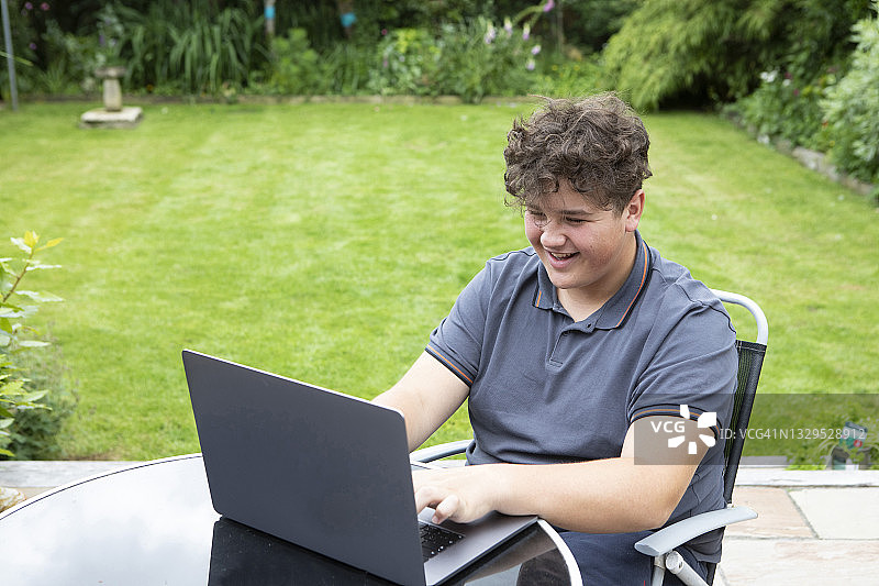 一个十几岁的男孩微笑着坐在花园的桌子旁使用笔记本电脑图片素材