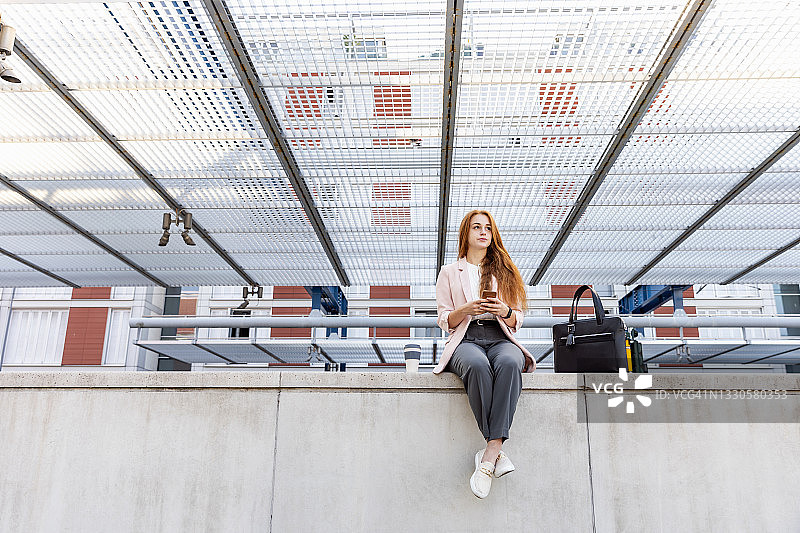 女自由职业者坐在火车站的挡土墙上，拿着手机看向别处图片素材