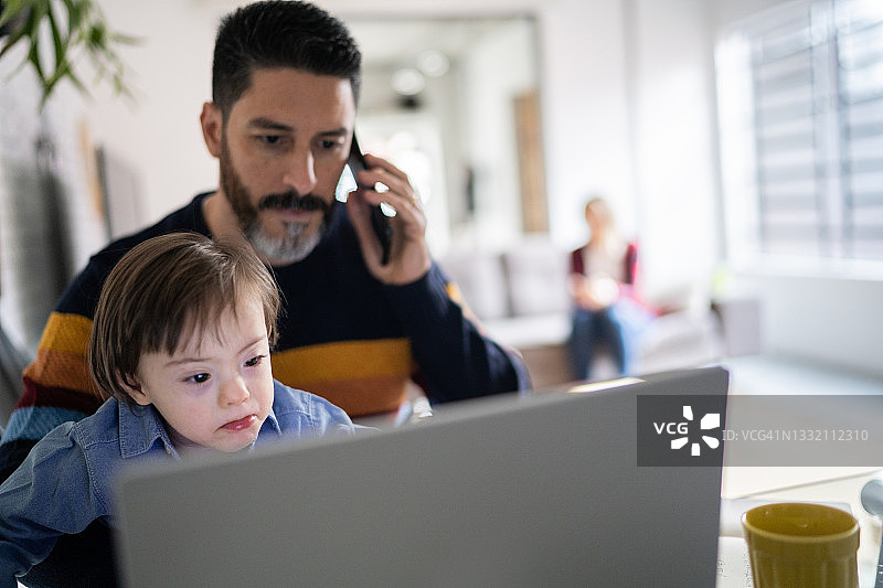 一个成熟的男人一边在家工作一边打电话，带着一个有特殊需要的儿子图片素材