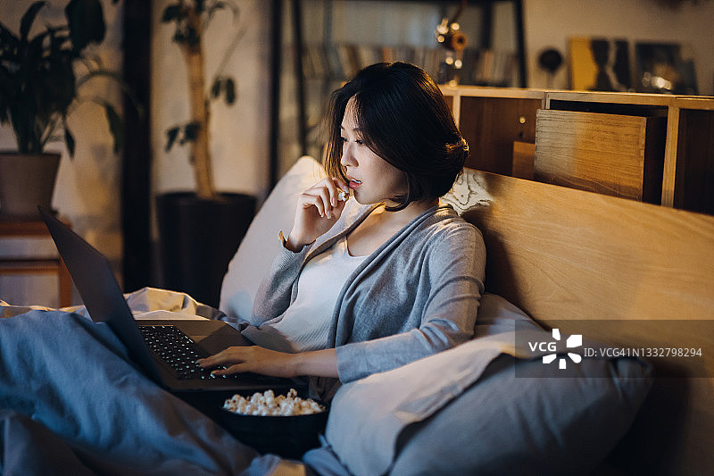 年轻的亚洲女人躺在床上享受周末，晚上在家看笔记本电脑上的电影和吃爆米花图片素材