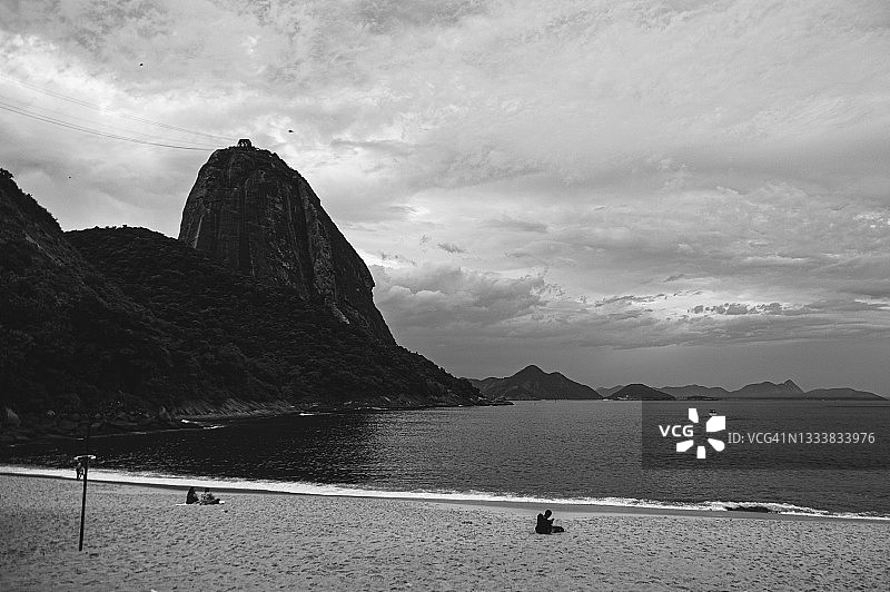 人们在红色海滩(Praia Vermelha)上放松，旁边是Pão de Açúcar (Sugarloaf)山，天空阴沉沉的图片素材
