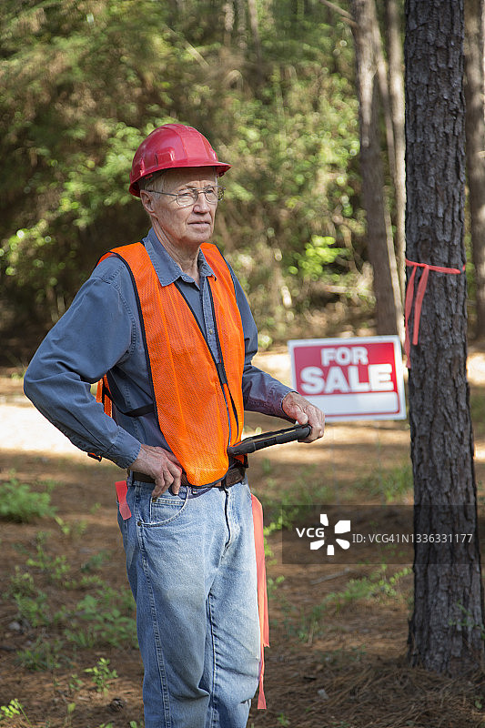 施工经理正在标记要砍伐的树木。他随身携带一台电子平板电脑，穿着安全背心，戴着安全帽。房屋上挂着出售标志。图片素材