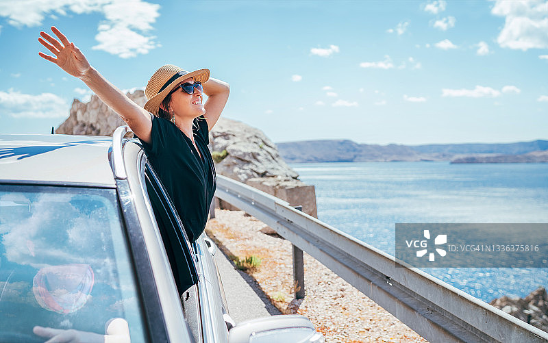 愉快的女人肖像享受海滨自驾游。她穿着黑色的衣服，戴着草帽和太阳镜，张开双臂，幸福得发亮。暑假汽车旅行的概念。图片素材