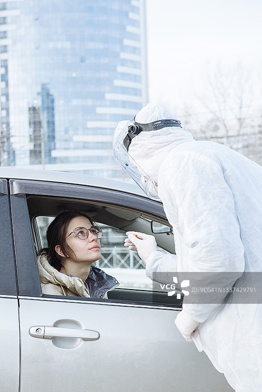 穿着个人防护用品防护服的病毒学家或医生用棉签从一名女司机的车中提取PCR检测样本。新型冠状病毒移动检测概念图片素材