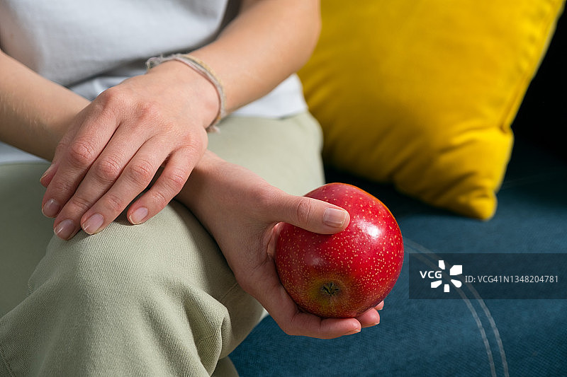 一个坐在家里色彩鲜艳的沙发上的女人手里拿着一个红苹果。一个女孩在吃水果。有机农产品的种植。素食、纯素、生食和饮食的概念。图片素材