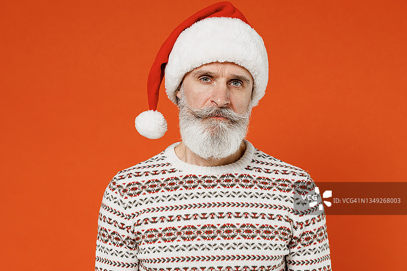 50多岁的圣诞老人戴着圣诞帽，穿着毛衣，摆着姿势，在橙色背景的摄影棚里孤立地看着镜头。祝2022年新年快乐，庆祝圣诞佳节快乐图片素材
