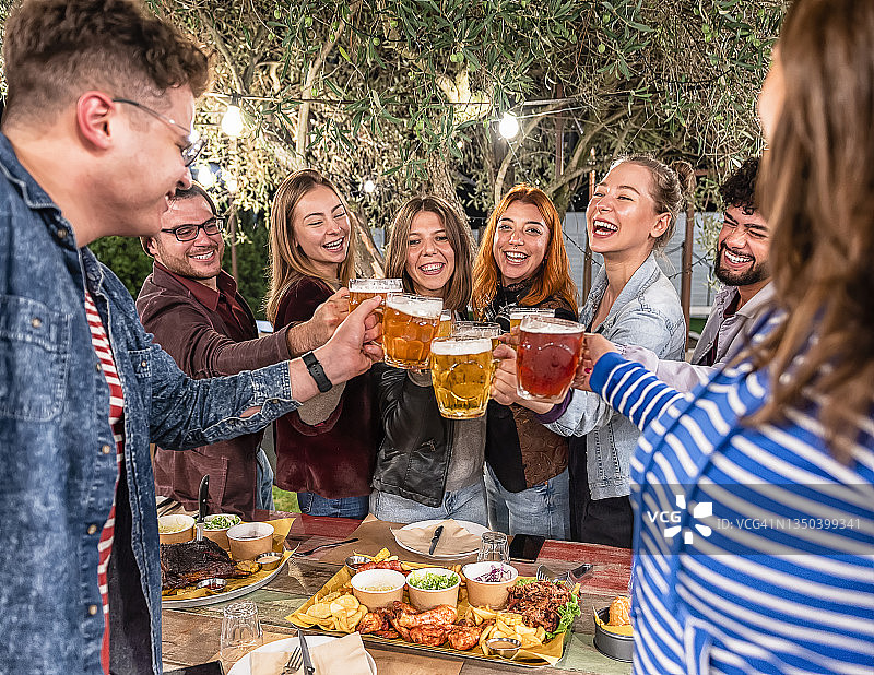 快乐的男孩和女孩在餐厅的花园中喝啤酒-一群朋友坐在夏天的露台上，手里拿着啤酒，桌上放着烧烤-友谊和青春的概念图片素材