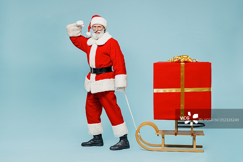 全身健壮的圣诞老人男子50多岁，戴着圣诞帽，身穿红色套装衣服，拉着圣诞雪橇，带着礼盒，在素色背景的演播室里展示肌肉。新年快乐，2022，圣诞快乐图片素材