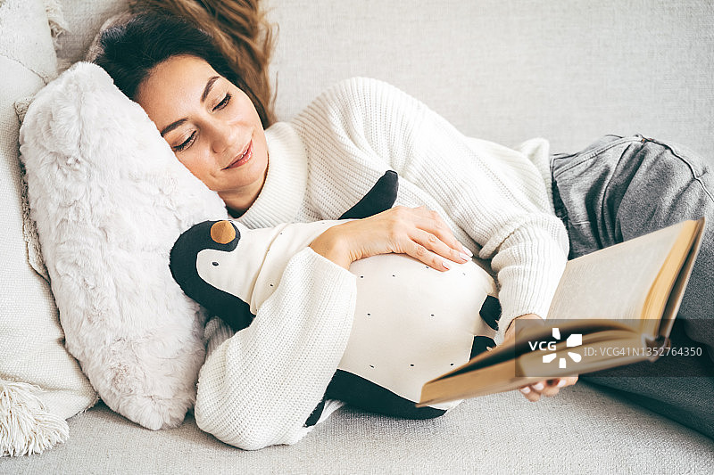 美丽的睡眼惺忪的女人正在舒适的沙发上看书，抱着她的毛绒企鹅玩具图片素材