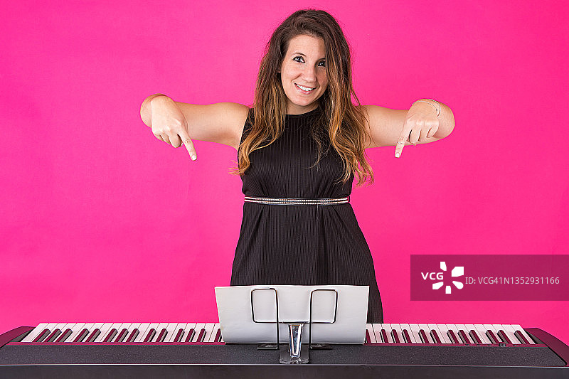 钢琴家音乐家女孩身穿黑色连衣裙，戴着一个带有高音谱号的戒指，手指指向钢琴，在粉红色的背景上。音乐，钢琴家和作曲家的概念。图片素材