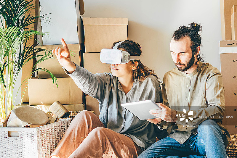 快乐的年轻夫妇规划室内设计在虚拟现实。一个男人拿着平板电脑，一个女人戴着现代VR头盔，坐在纸板箱中间，与增强现实的物体一起工作。图片素材