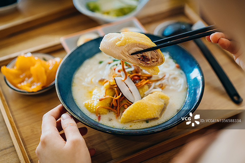 从肩膀上看，一名亚洲妇女在餐厅用筷子吃着一碗新鲜的鸡肉和蔬菜汤面，盛在木托盘上。亚洲饮食文化图片素材