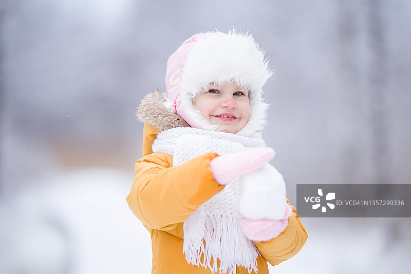 快乐的微笑2岁幼儿在寒冷的日子里做和拿雪球。一个小女孩戴着毛皮帽子，戴着白色围巾，戴着手套，穿着黄色温暖的夹克。特写镜头。前视图。图片素材