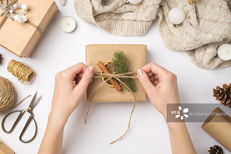 第一个人俯视图照片的女性双手绑绳蝴蝶结礼品盒与松枝肉桂棒干燥柠檬片围巾圣诞树球和工艺品在孤立的白色背景图片素材