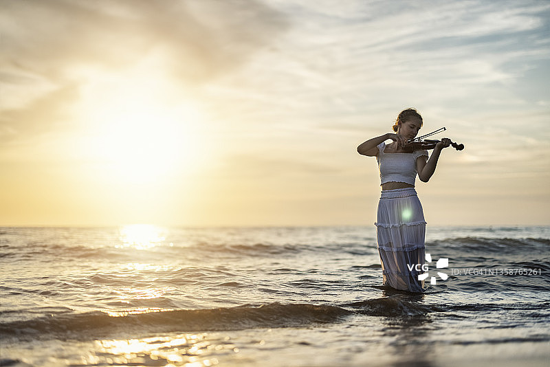 一个十几岁的女孩在日落的海滩上拉小提琴图片素材