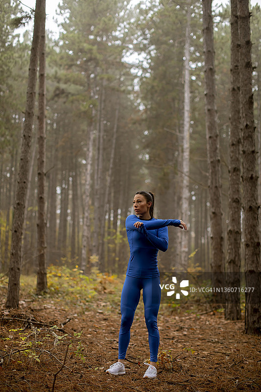 在秋天的森林里，一名身着蓝色运动服的年轻女子在锻炼前伸展身体图片素材