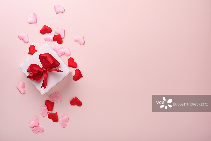 白色的礼盒，红色的丝带和小的红色装饰心在粉红色的背景上。顶部视图与复制空间。情人节或婚礼的浪漫概念。节日的作文。模拟。图片素材