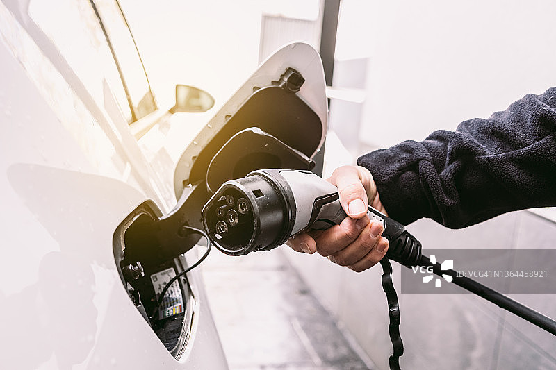 详细介绍:一只手将电线插进一辆电动汽车，在车库外为电池充电。电动汽车充电、可再生能源、可持续性和交通的概念。图片素材