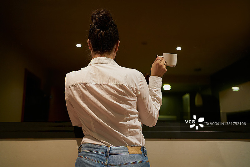 女性挽着发髻，手拿咖啡，透过玻璃向外看图片素材