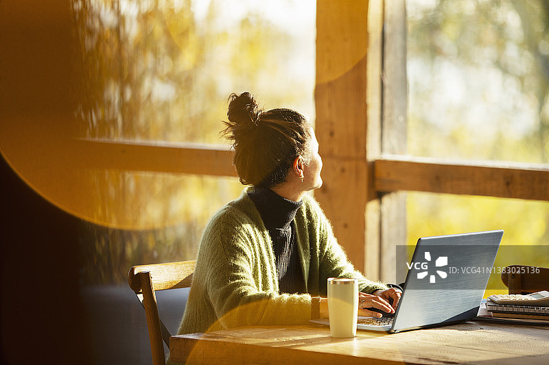在阳光明媚的咖啡馆里，坐在笔记本电脑前看着窗外的女人图片素材