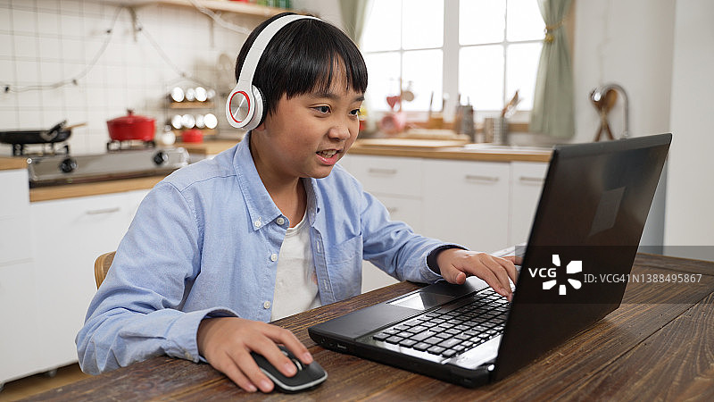 可爱的亚洲小男孩，戴着耳机坐在家里餐厅的个人电脑上玩游戏图片素材