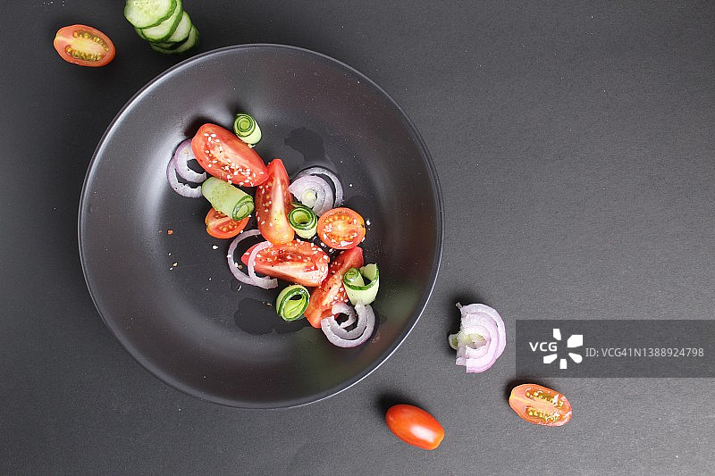 蔬菜沙拉，用西红柿、黄瓜、红青瓜和白芝麻种子放在黑色的盘子里，背景是黑色的。健康的食物健康的生活方式图片素材