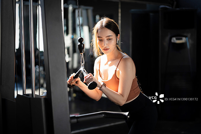 图为在健身房的机器上锻炼的年轻亚洲女性的肖像。快乐的肌肉健美女性在健身中心锻炼，通过在线健身教练使用笔记本电脑进行训练图片素材