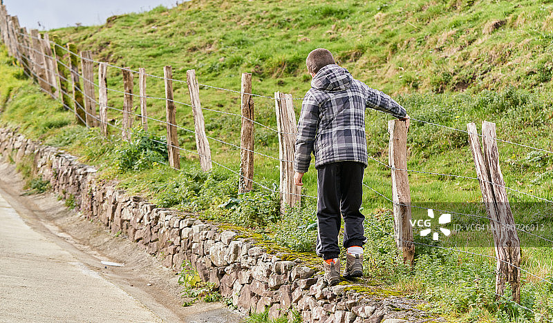 在一个寒冷的冬日，一个男孩穿着暖和的衣服，走在乡间小路的石头边，紧紧地抓住一个私人农场的铁丝围栏。图片素材