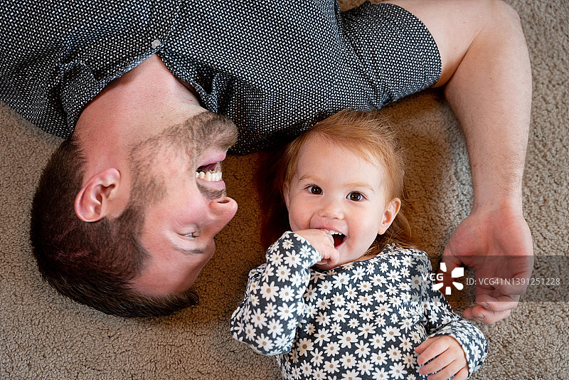 年轻的父亲和他可爱的小女儿躺在地板上，有爸爸，女儿结合时间，微笑，大笑，看着对方图片素材
