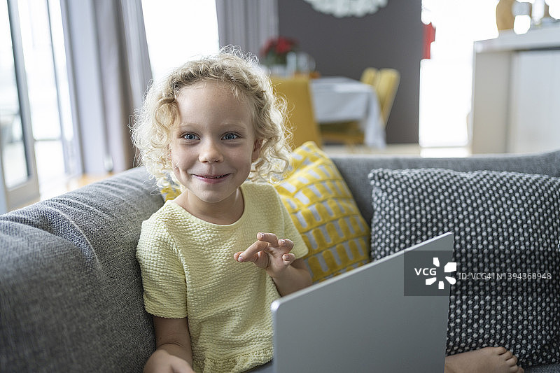 快乐的金发女孩带着笔记本电脑坐在家里客厅的沙发上图片素材