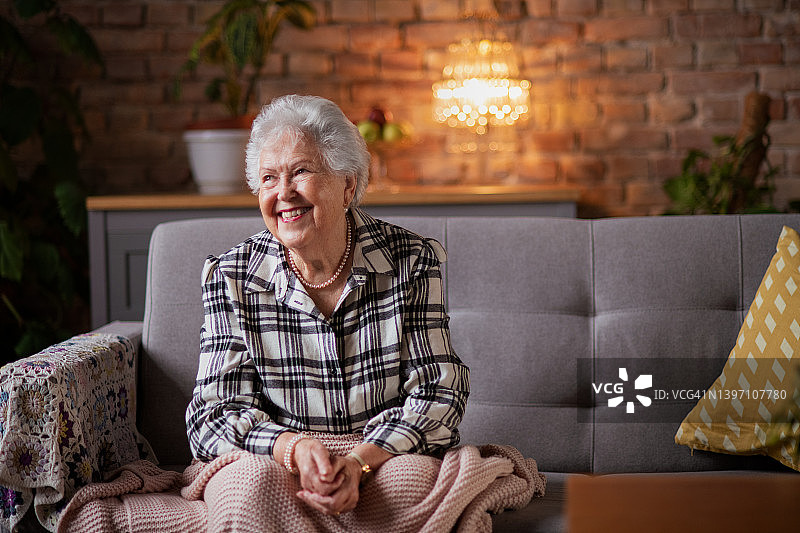 快乐的老妇人裹着毯子坐在家里的沙发上。图片素材
