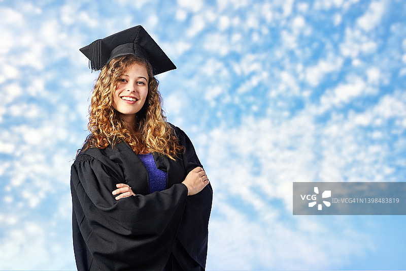 自信而自豪的女学生抱臂站着，戴着毕业帽，穿着毕业服，微笑着图片素材