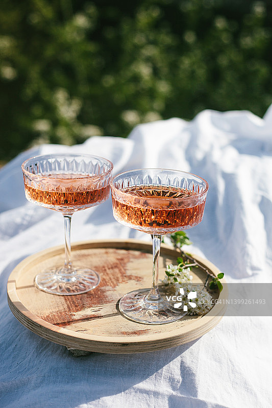 两杯玫瑰红葡萄酒放在户外的木托盘和白色的纺织品，在春天野餐。阳光明媚的日子，绿草如茵，樱花盛开。图片素材