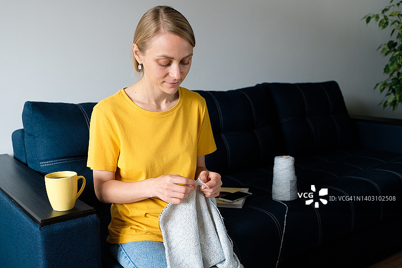 女孩手里拿着一件产品，正在织针。旁边有一杯咖啡或茶。一个女人坐在家里的沙发上编织羊毛衫或围巾。爱好、创意、针线活和手工工作的概念。副本的空间。图片素材