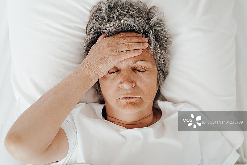 俯视成熟疲惫的女人闭着眼睛抱着头躺在床上的枕头上。失眠、头痛、压力、抑郁的概念图片素材