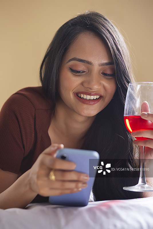 年轻女子在床上用智能手机端着一杯酒图片素材