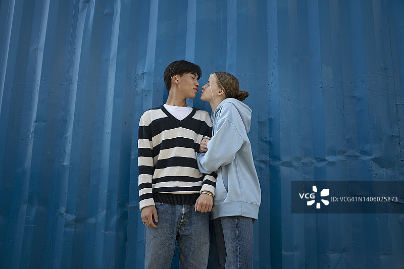 十几岁的男孩和女孩靠在一个蓝色的金属工业容器上，面对着对方，抿着嘴唇。图片素材