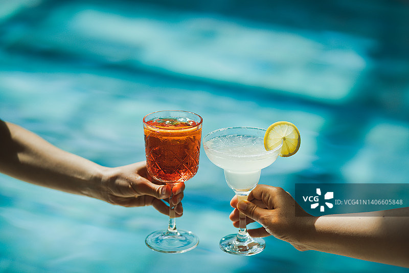 在游泳池背景下，两只手拿着阿佩罗和玛格丽塔鸡尾酒举杯。干杯图片素材