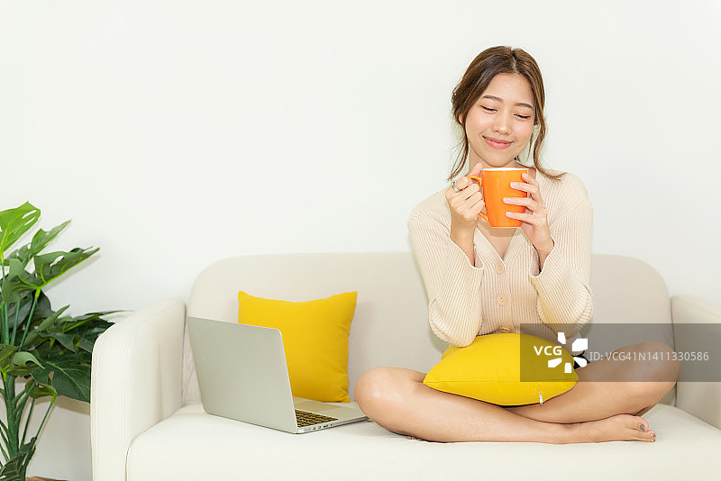 快乐的年轻女子喝热橙杯咖啡后，工作和网上会议在家里。自由自在的生活方式，在客厅放松和放松。生活方式的概念图片素材