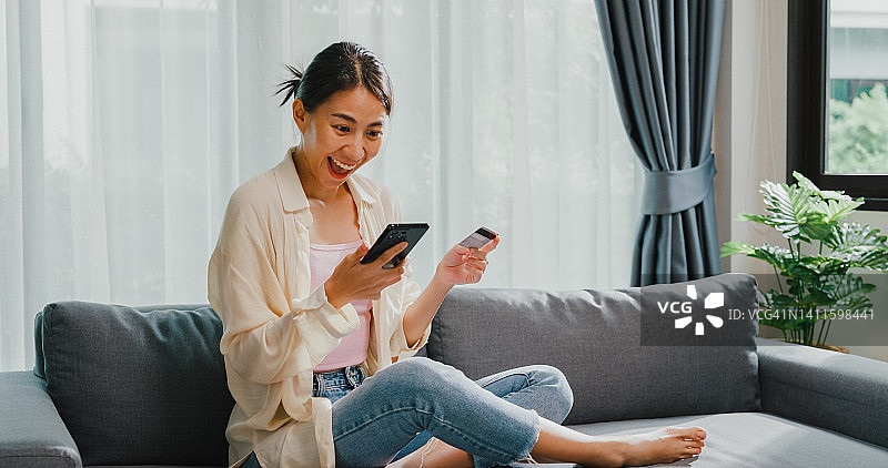 年轻的亚洲女女孩穿着休闲坐在沙发上，抱着智能手机和信用卡感觉兴奋和幸福的网上购物成功支付在客厅在家里。图片素材