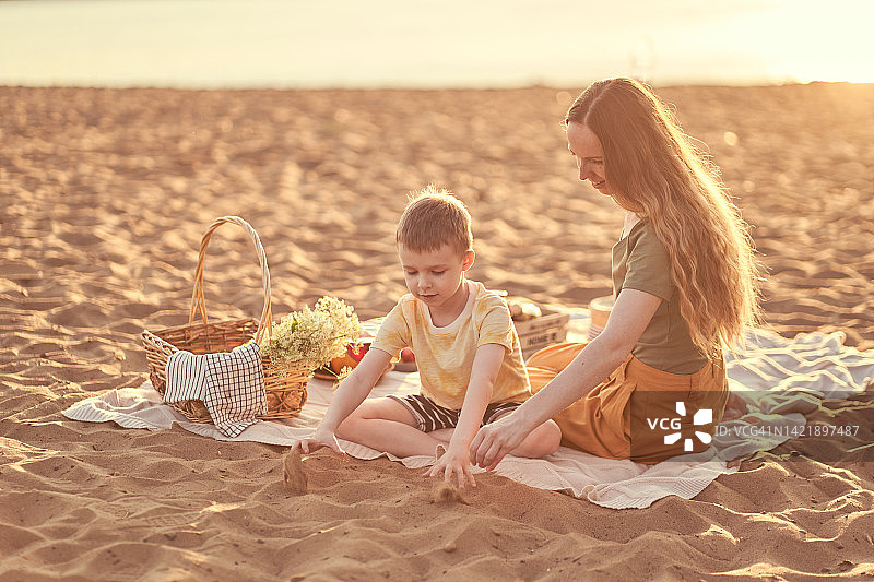 沙滩上一个美丽的女人和她的儿子坐在野餐毯子上玩沙子。一个年轻的女人带着一个孩子在海滩上野餐，篮子上放着食物和饮料。图片素材