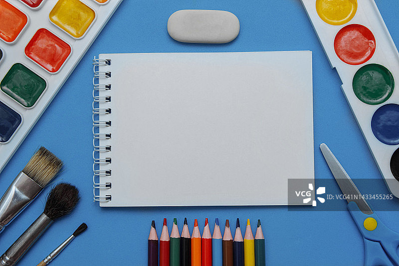 水彩颜料，刷子，铅笔，剪刀，铅笔和白色记事本在一个蓝色的背景和空白的文本。图片素材