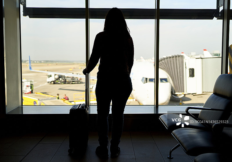 在灯光的映衬下，一个长发女人，拿着她的行李放在窗前，注视着机场准备登机的飞机图片素材