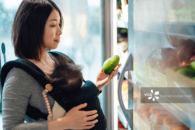年轻妇女在超市买有机蔬菜，她抱着她的婴儿背带的女儿图片素材