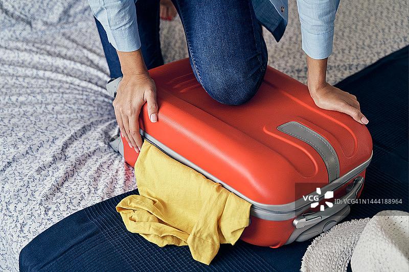 特写镜头，一个不认识的女人爬上一个装满衣服的行李箱，试图关闭它，去度假。图片素材