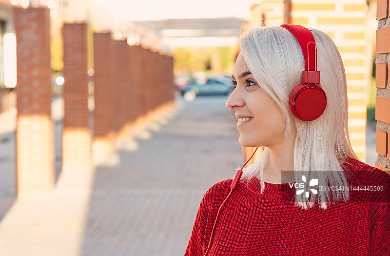 银发女孩戴着红色耳机听音乐。靠在城里的一根柱子上。穿着一件红毛衣。图片素材