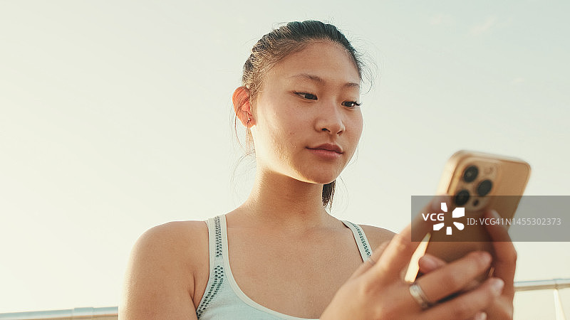 在晨光中，站在现代建筑背景的海滨，微笑的亚洲女孩在运动上衣使用手机图片素材